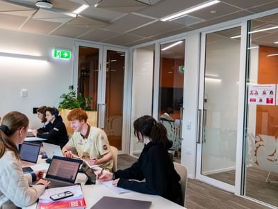 Students collaborating at tables with laptops in a modern meeting space with glass-walled conference rooms at Tupuārangi.