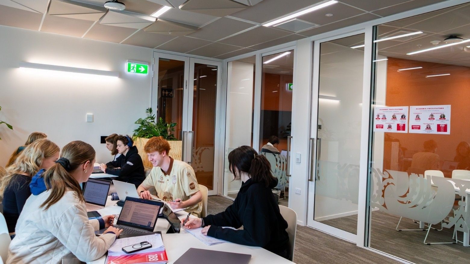 Students collaborating at tables with laptops in a modern meeting space with glass-walled conference rooms at Tupuārangi.