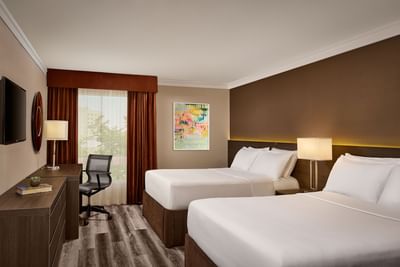 Double guest room with two beds, nightstands, artwork, desk, chair, TV, and window with curtains.