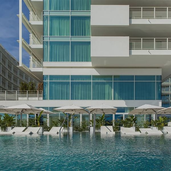 Modernes Gebäude mit Pool, Liegestühlen und Sonnenschirmen vor blauem Himmel.