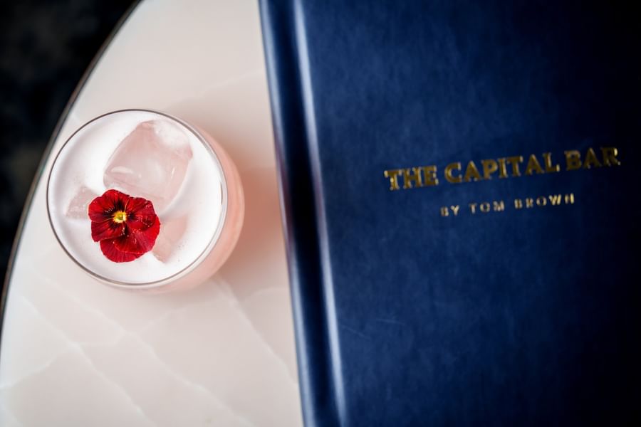 Pink cocktail with a red flower garnish placed by a blue menu at The Capital Hotel, Apartments and Townhouse