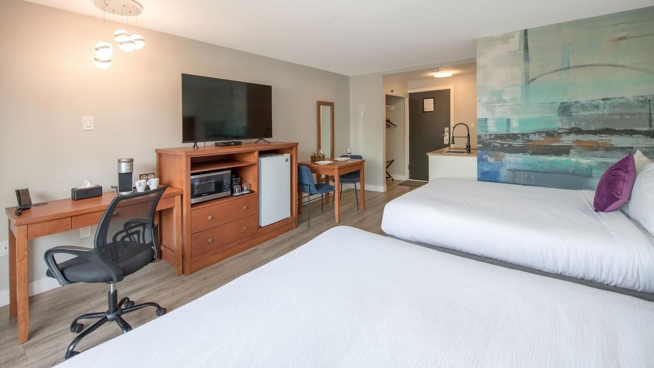 Two queen beds, desk with chair, TV, mini fridge and small kitchen area in a hotel room.