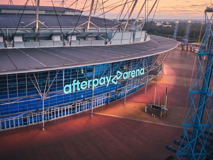 Exterior view of Afterpay Arena with illuminated blue signage at dusk, showing modern glass facade and surrounding plaza.