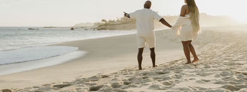 A couple holding hands walks along a sandy beach at sunset, enjoying a serene moment near Marquis Los Cabos Resort