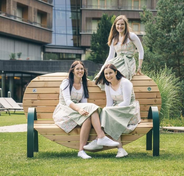 Tre donne in abiti tradizionali sedute su una panchina di legno davanti a un edificio.