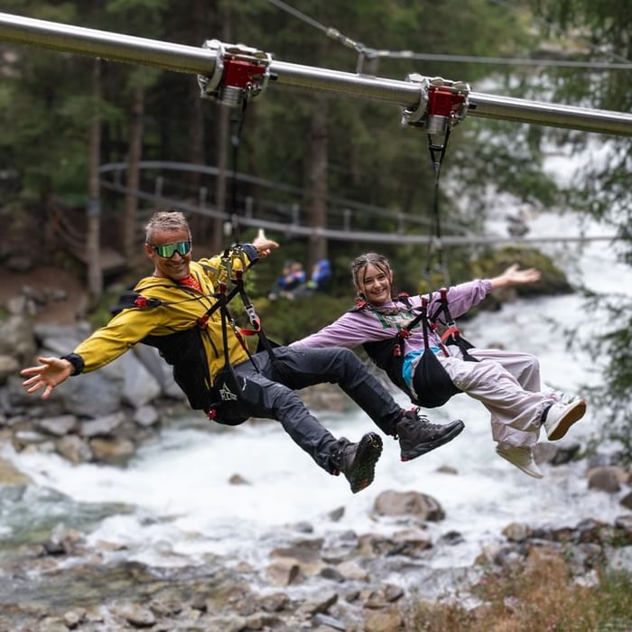 Uomo e donna sorridenti in parapendio, sospesi sopra un fiume di montagna.