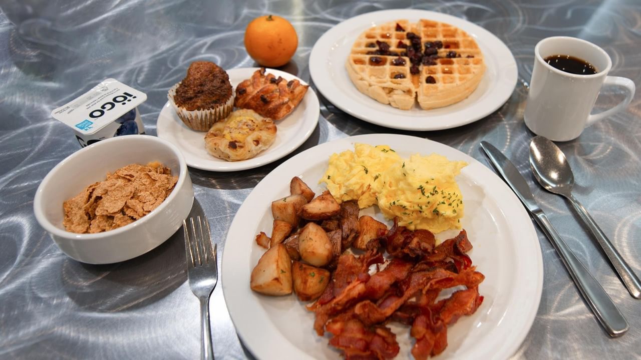 Breakfast spread with scrambled eggs, bacon, potatoes, waffles, muffin, pastries, cereal, orange, and coffee on a bed tray.