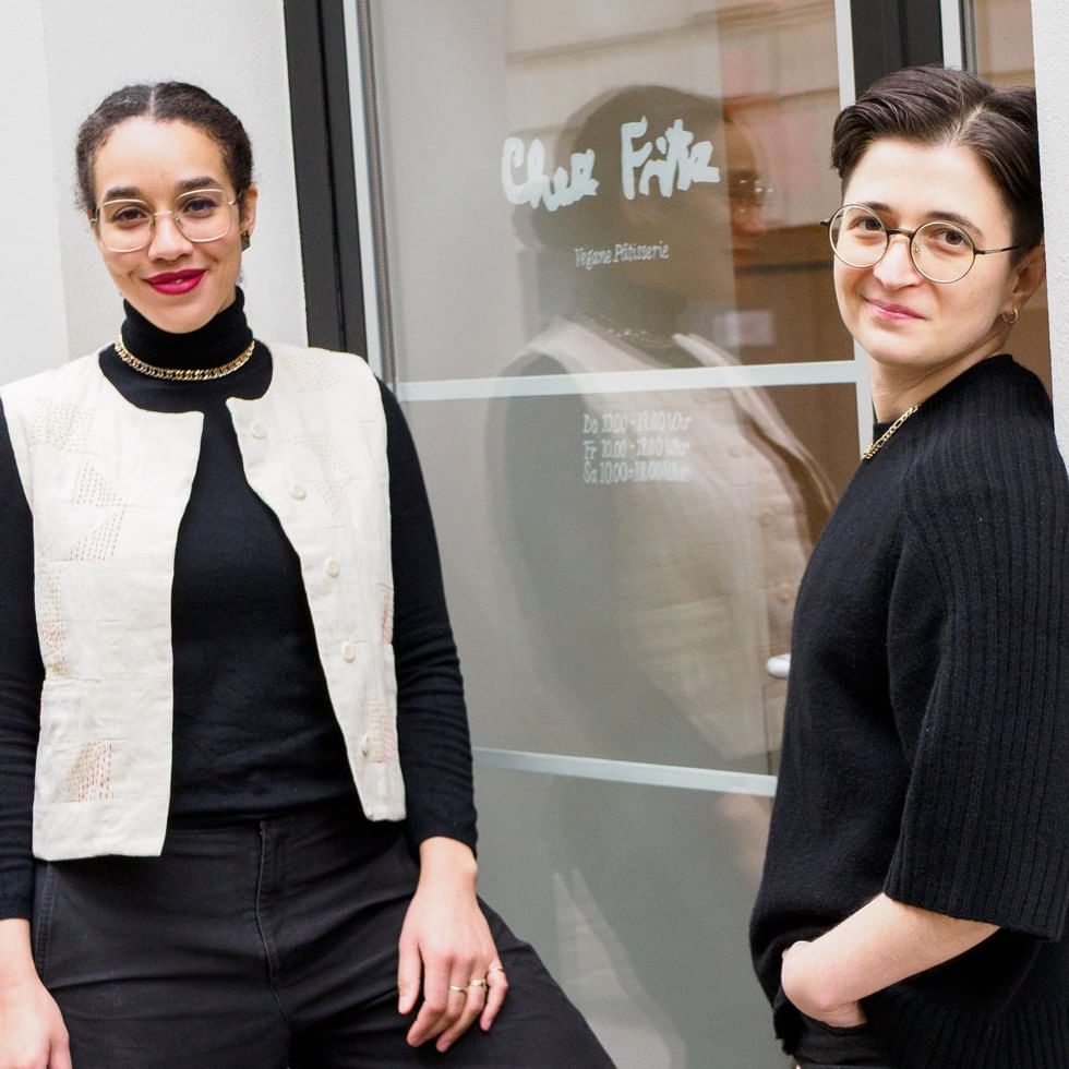 Two women wearing glasses stand in front of the Chez Fritz vegan patisserie storefront window in Vienna.
