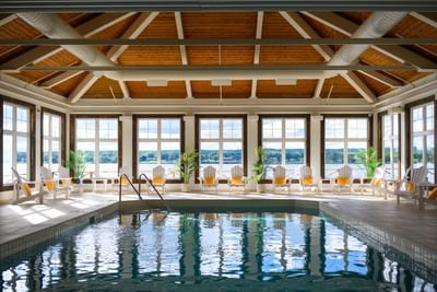 Indoor pool by white lounge chairs under a high wooden roof, with scenic lake views at Rodd Miramichi River