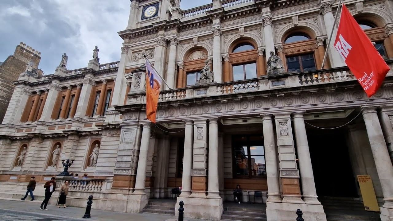 The historic grand stone facade of the Royal Academy of Arts building displaying red flags near The Londoner Hotel
