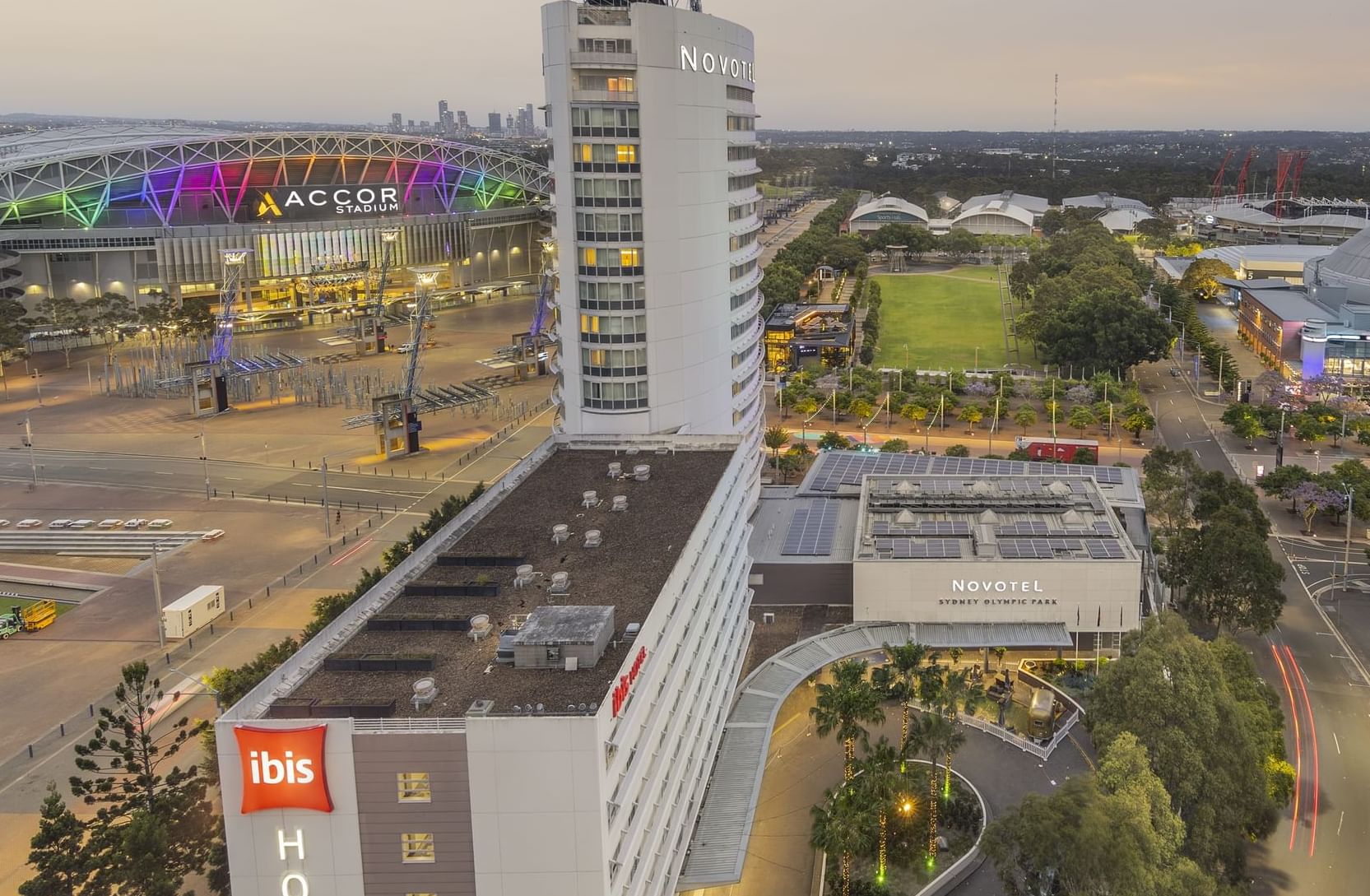 Aerial view of Novotel Sydney Olympic Park, featuring the Driveway, Accor Stadium beside it & the city in the background