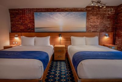 Two beds between side tables against an exposed brick wall with sunset art in Double Queen City View at Harborside Inn Boston