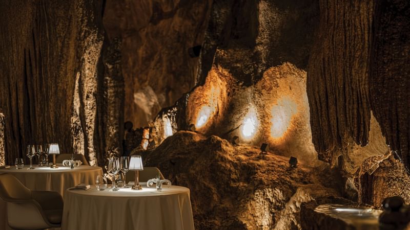 Two tables with white tablecloths, wine glasses, and lit lamps in a cave setting with lighting.