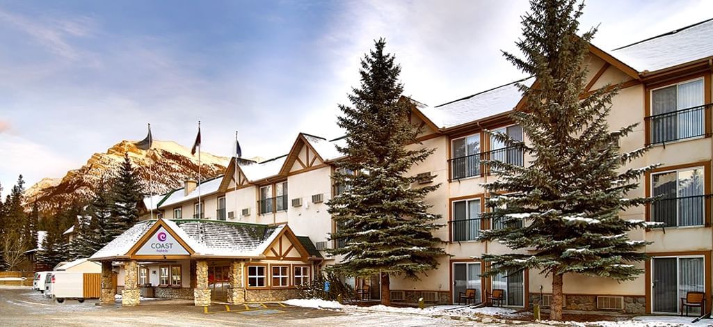 A scenic view of Coast Canmore Hotel & Conference Centre, featuring a charming exterior with snow-covered trees and mountains in the background, creating a cozy and inviting winter atmosphere.