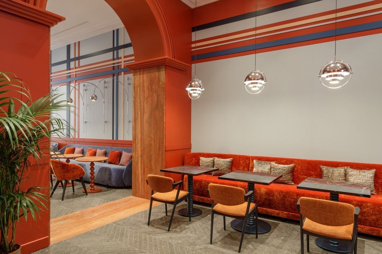 Stylish dining area at the Independent Hotel in Rome with long banquette seating, velvet chairs in deep blue and purple tones, marble tables, red and orange accents, and modern chrome wall lamps under high ceilings.
