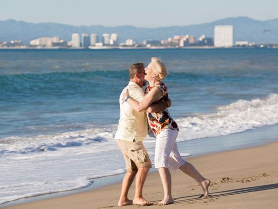 Joyful senior couple embraces and laughs on a sandy beach with a city skyline behind them near Playa Los Arcos Resort & Spa