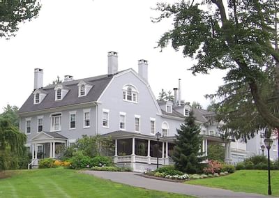 Exterior view of Simsbury 1820 House, a gambrel-roofed mansion with a wraparound porch surrounded by gardens and lawns