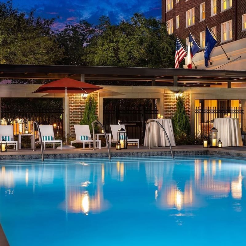 Illuminated pool Side by lounge chairs under a red umbrella near a brick building at Warwick Melrose Dallas