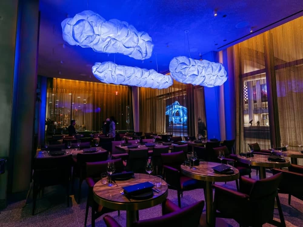 a dimly lit dining room with round tables and chairs, large windows, and unique cloud-shaped lighting fixtures