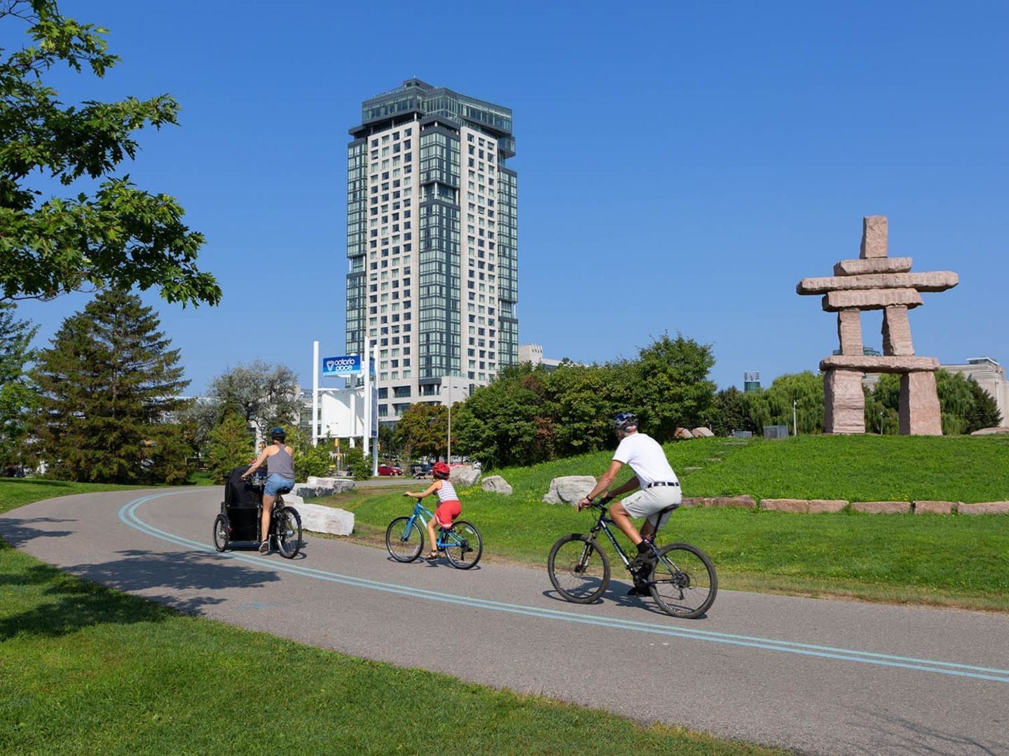 Cyclists riding a paved lakeside path by the stone Inukshuk statue with Hotel X Toronto tower rising above the trees