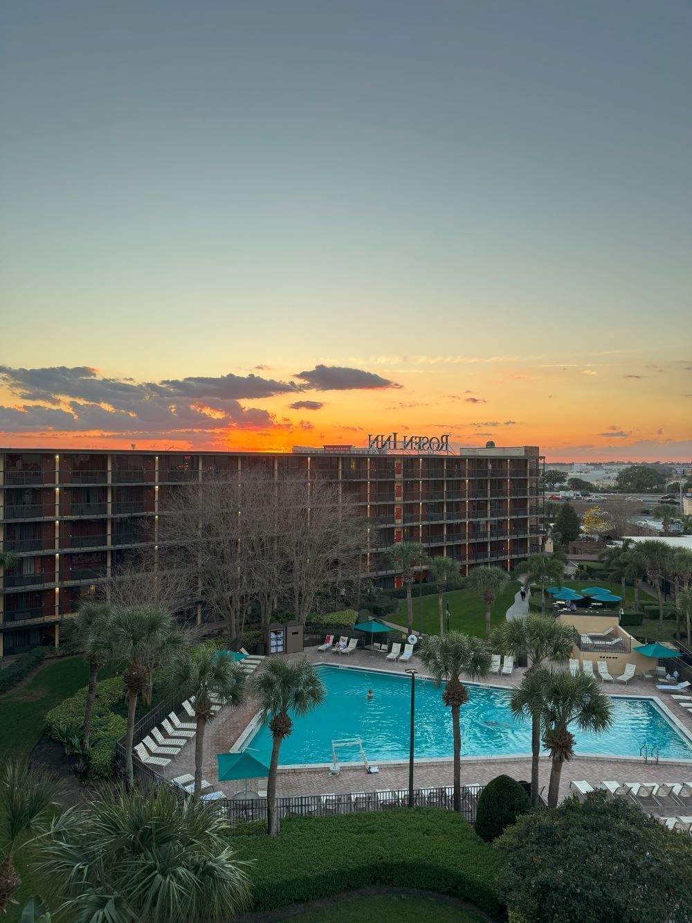 An elevated side view shows the multi-story Rosen Inn International hotel beside its large, bright blue swimming pool lined with palm trees and lounge chairs with a golden, orange sunset sky.