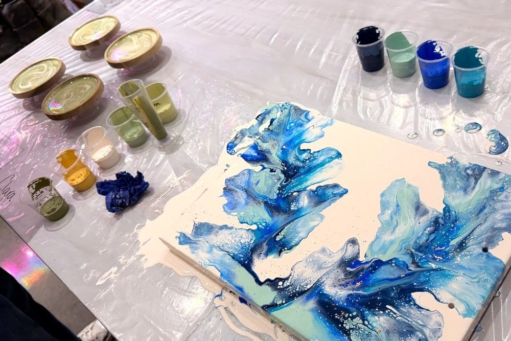 A large canvas with blue fluid art and four coasters with green fluid paint sit on a table beside cups of colorful paint at Abstract Orlando.