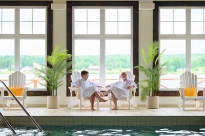 Two guests in robes relaxing by an indoor pool under tall windows, with green plants at Rodd Miramichi River