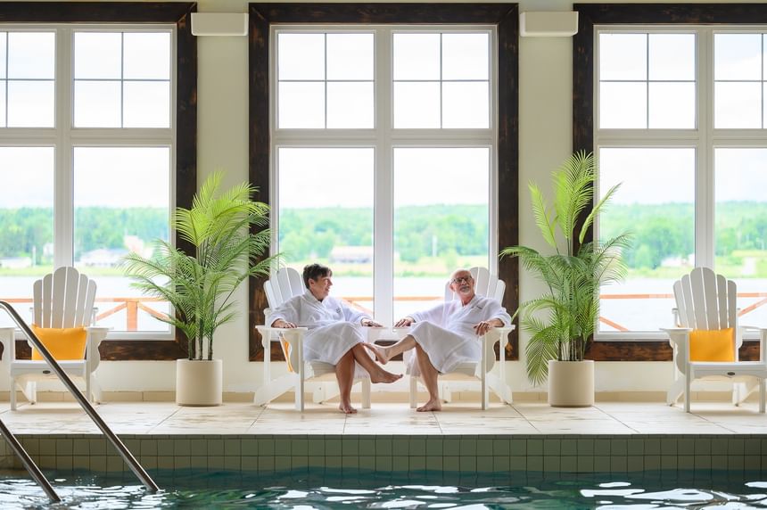 Two guests in robes relaxing by an indoor pool under tall windows, with green plants at Rodd Miramichi River