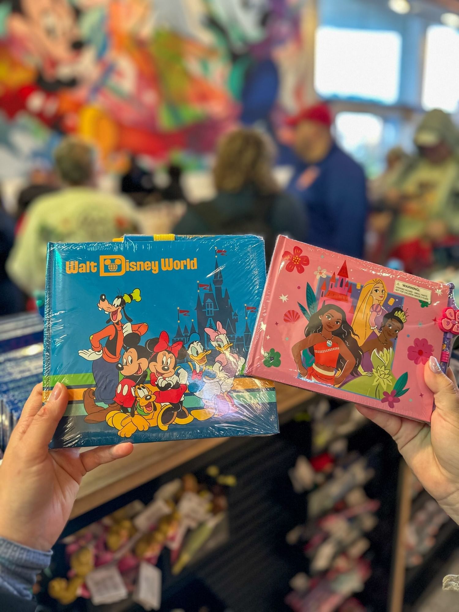 Two hands holding a blue Disney autograph book and a pink princess autograph book in an out of focus store.