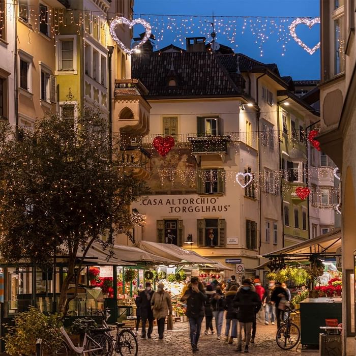 Strada cittadina decorata con luci e cuori per le festività, con negozi e persone che passeggiano.