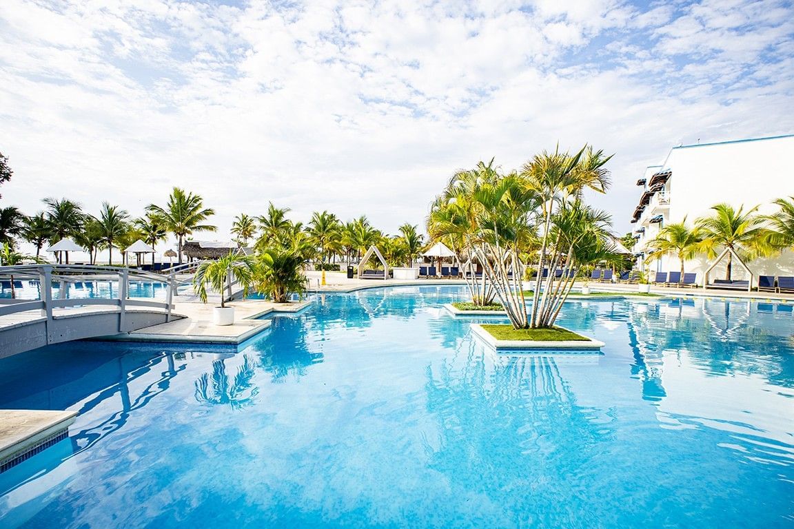 Large outdoor pool with island planters featuring palm trees and blue lounge chairs at Playa Blanca Beach Resort