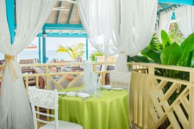 A romantic seaside dining setup featuring a green tablecloth, elegant tableware, and white drapes at Bay Gardens Inn