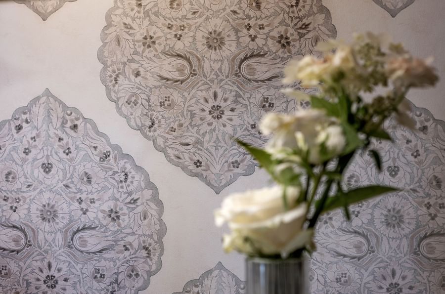 White flowers in a vase against a wall with intricate, paisley-inspired wallpaper design.
