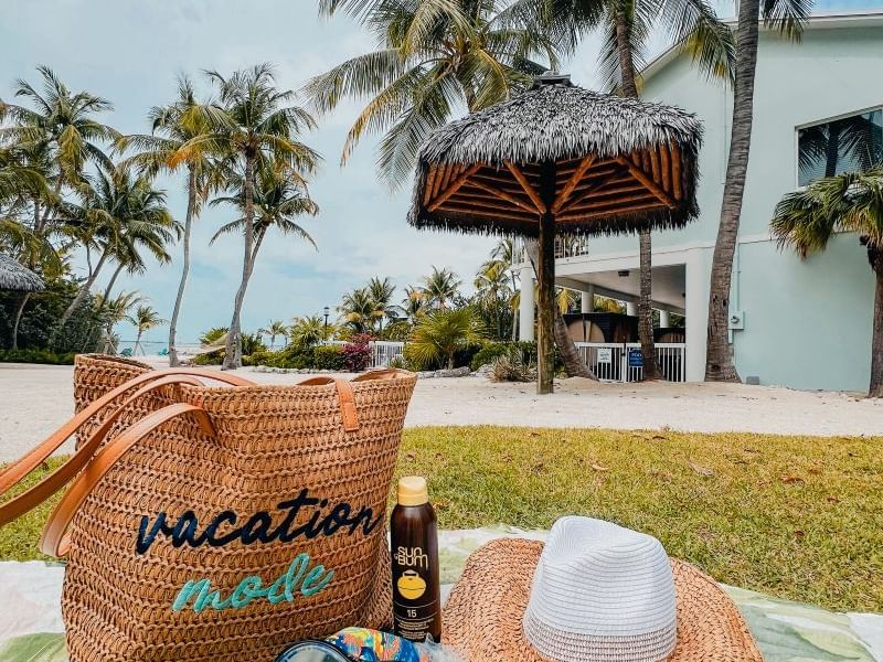 Vacation mode bag, snorkel mask, and straw hat on beach blanket near palm trees and resort.