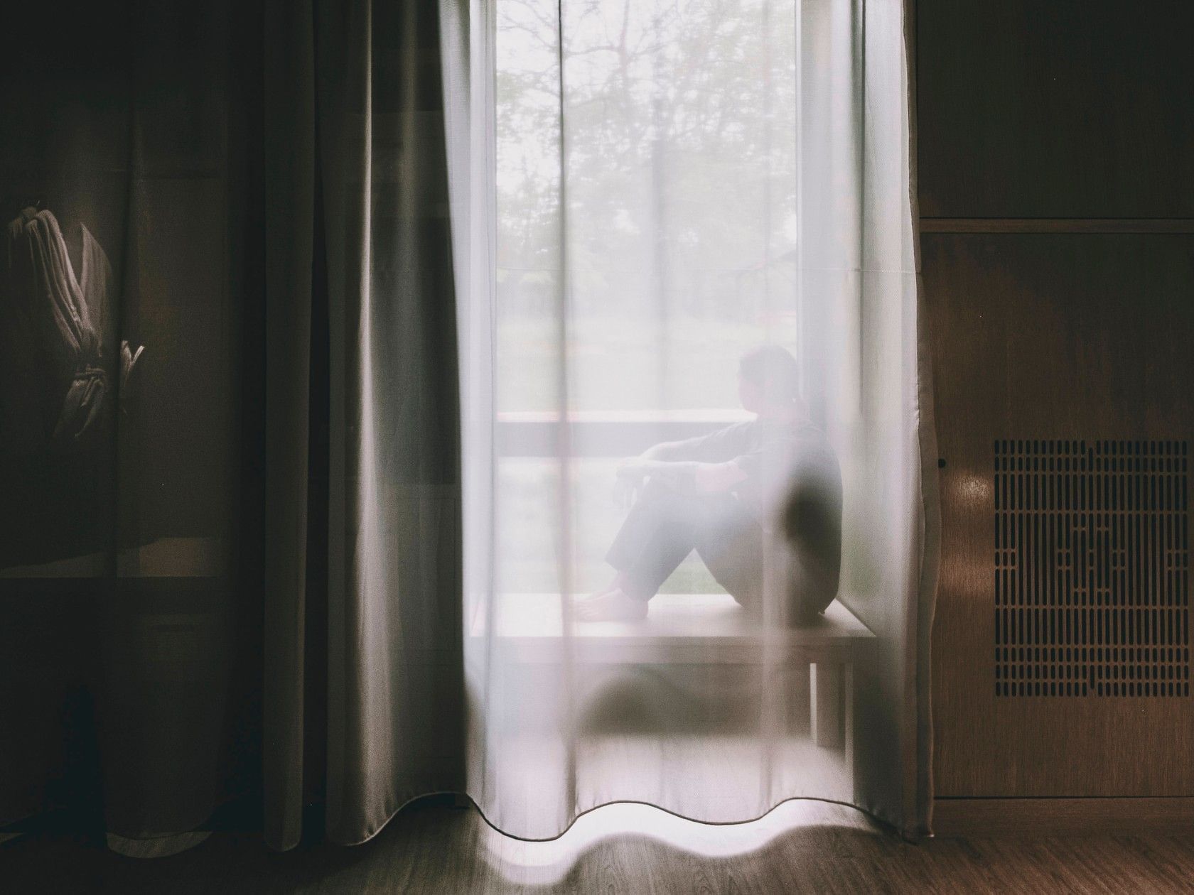 Person sitting on a window bench, visible through sheer curtains, with trees outside.