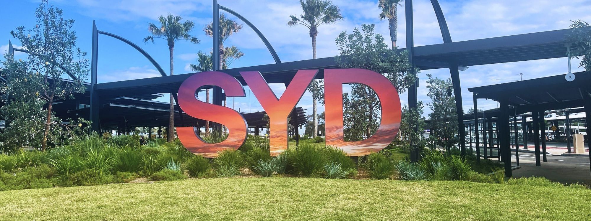 Vibrant SYD sign surrounded by greenery and palms near Novotel Sydney International Airport