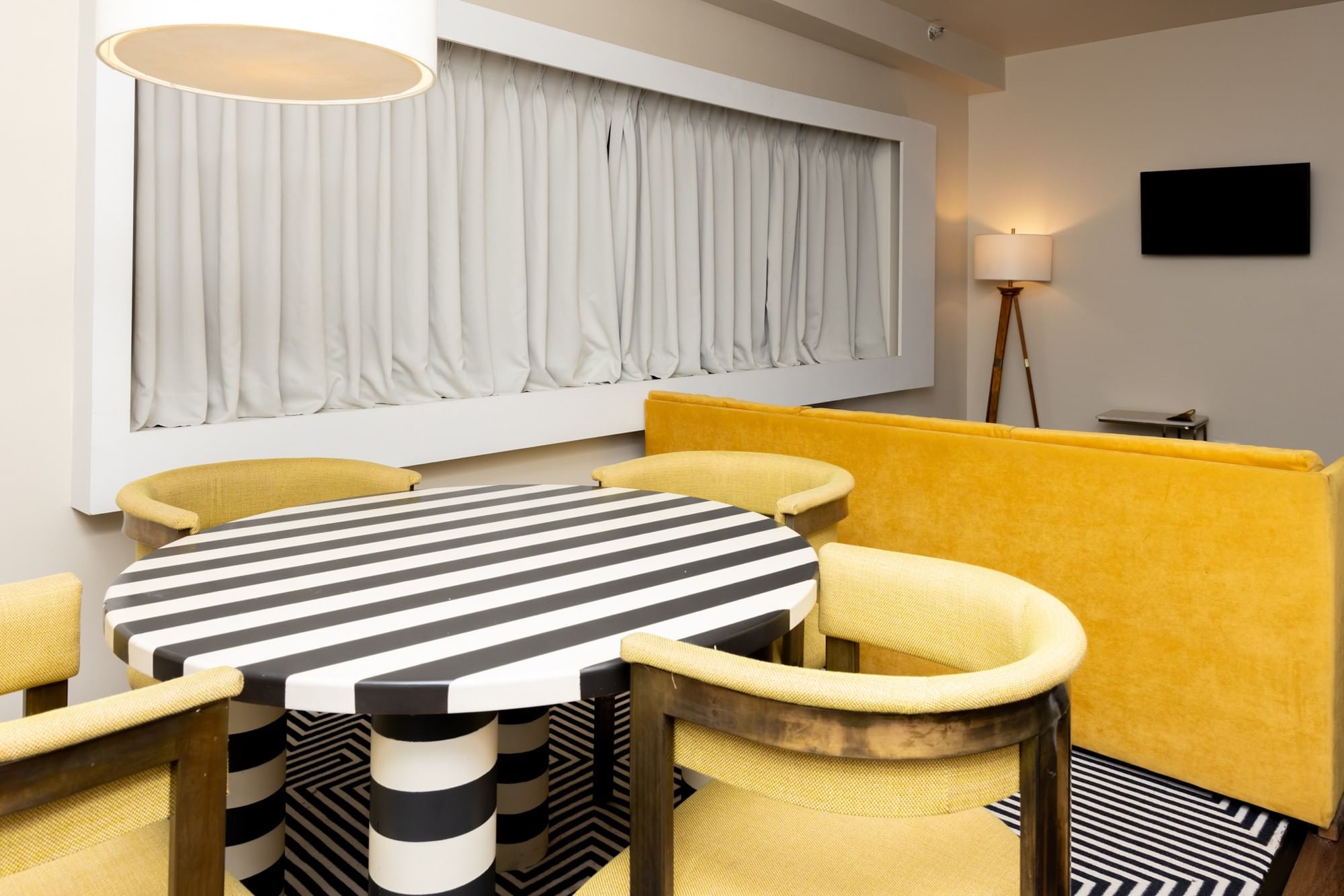 A dining area with a black and white striped table, yellow chairs and booth seating, a TV on the wall, and a lamp.
