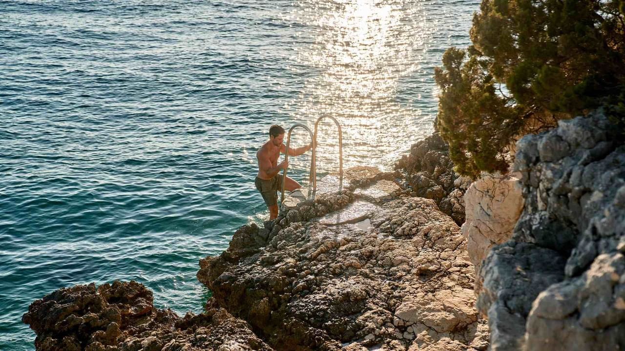 Uomo in costume, in piedi su roccia vicino al mare, che si appoggia a una ringhiera.