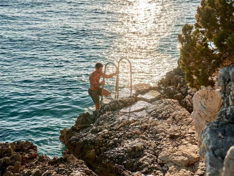 Mann in Badehose steht auf Felsen neben Meer, hält Metallstange und schaut in Wasser.