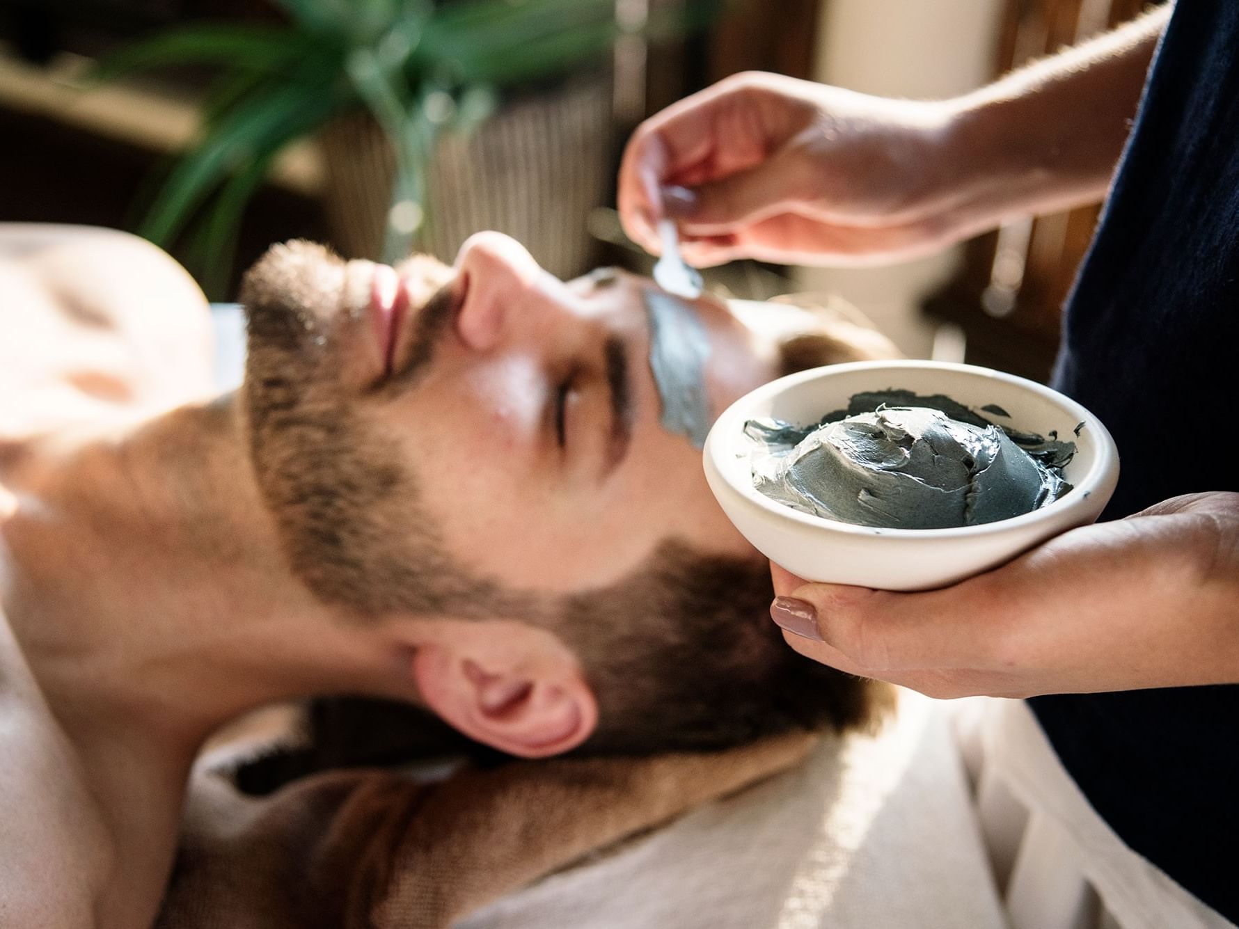 A man receives a facial treatment by a therapist, using a spatula and a bowl of product.