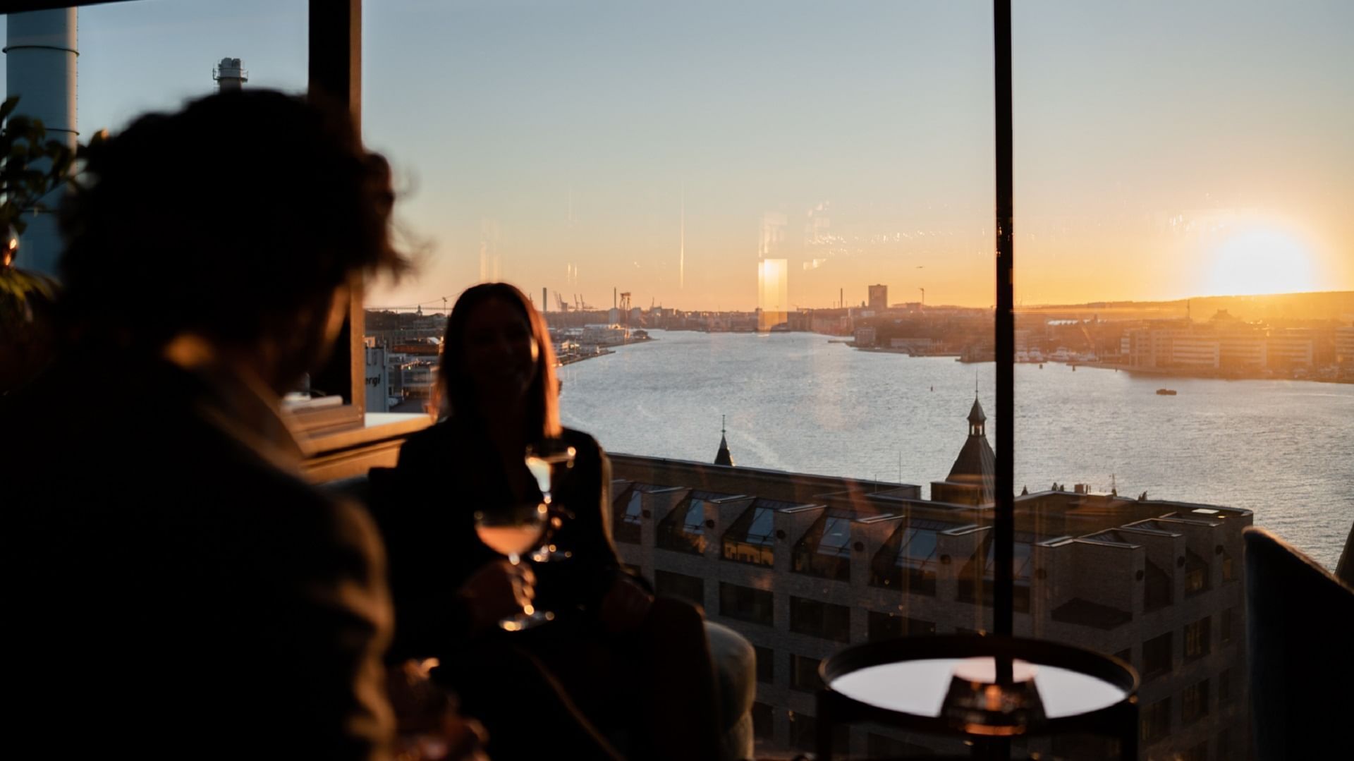 Par njuter av solnedgången över vatten med en drink i en modern stadsmiljö.