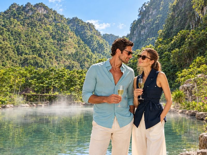 Couple holding champagne glasses by a misty hot spring pool surrounded by ancient limestone hills and greenery at The Banjaran Hotsprings Retreat
