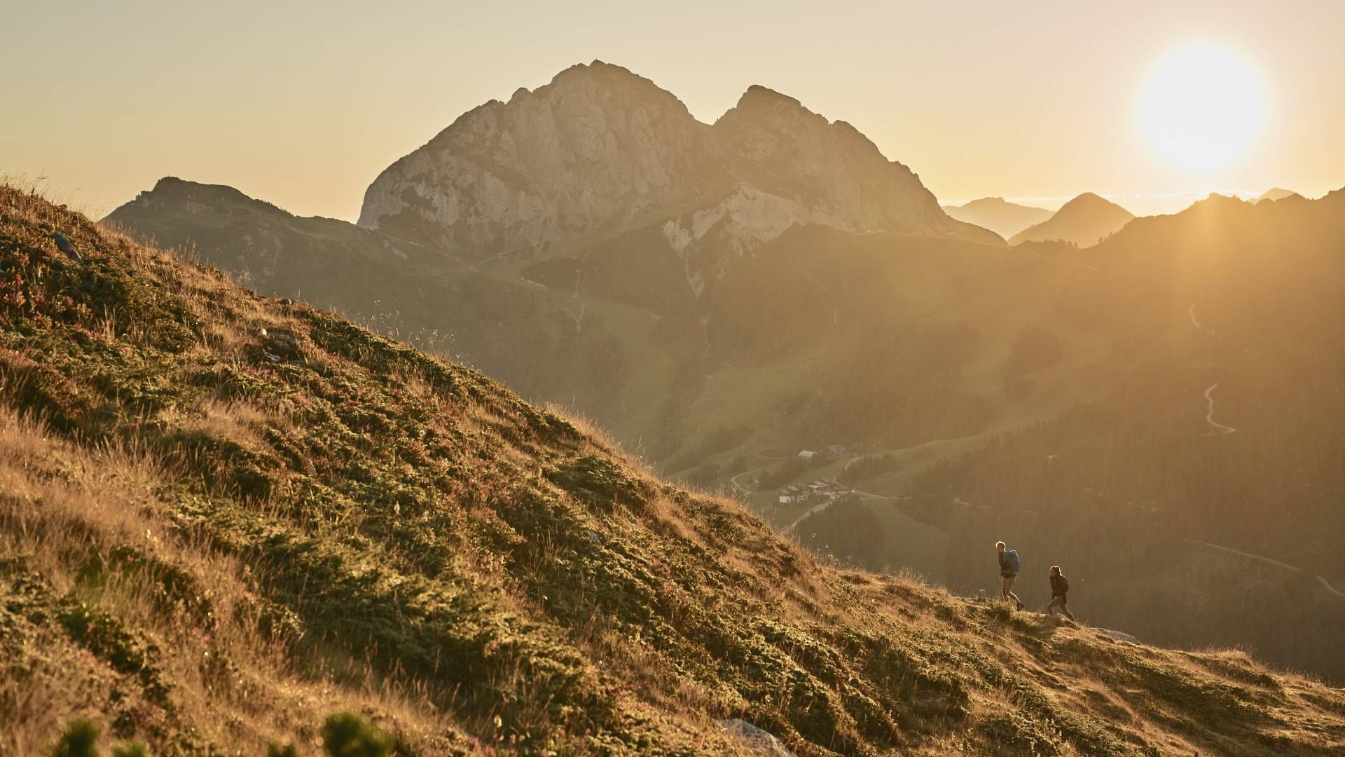 Dva planinara idu prema vrhu planine u sumrak dok sunce zalazi iza gorja.