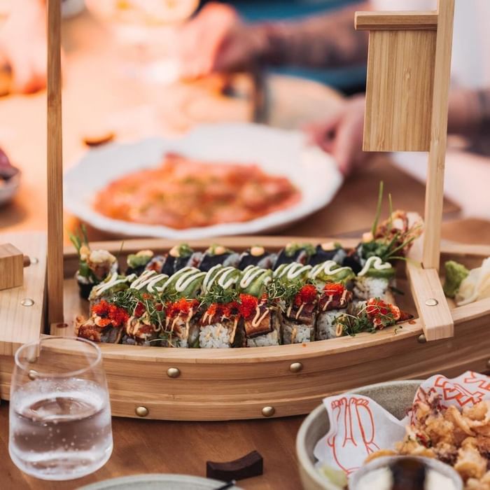 Sushi-Boot aus Holz mit verschiedenen Sushi-Rollen, einem Glas Wasser und weiteren Gerichten auf dem Tisch.