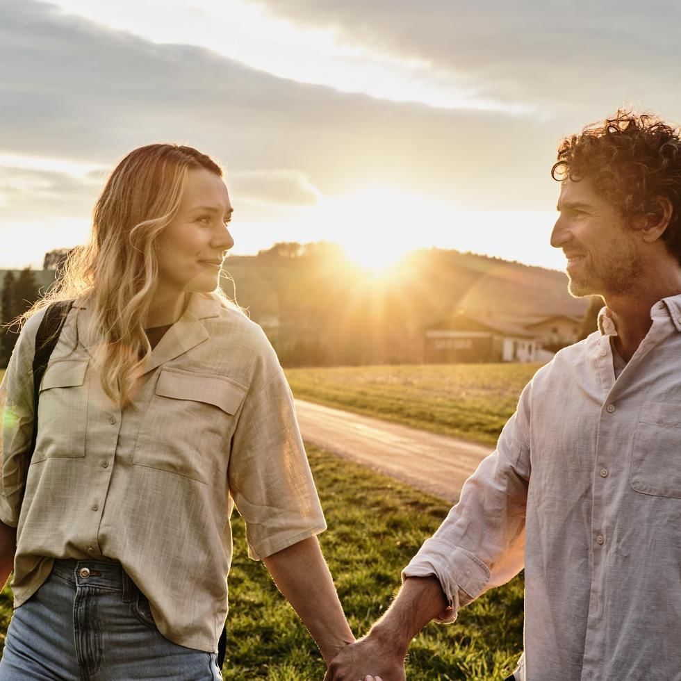 Man and woman holding hands walking down a dirt road in a field at sunset.