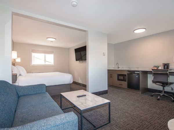 Modern hotel suite with a bed, couch, coffee table, and a small kitchenette.