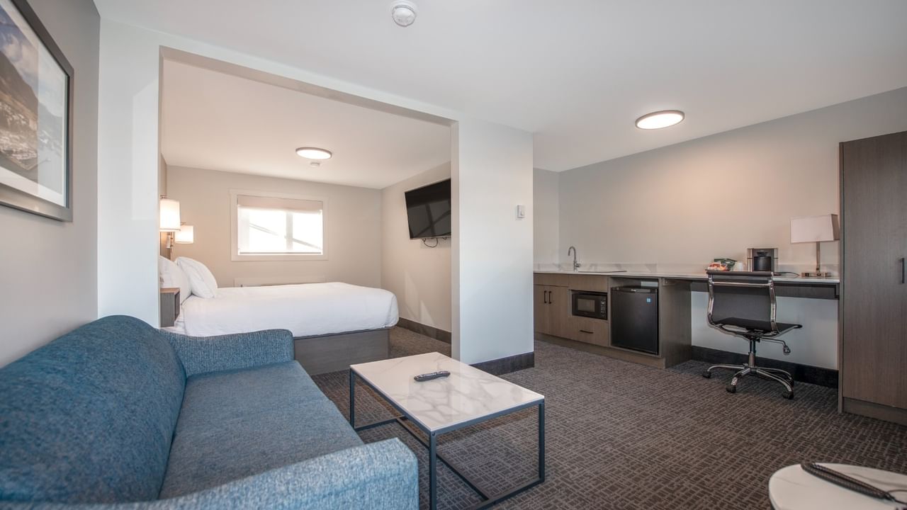 Modern hotel suite with a bed, couch, coffee table, and a small kitchenette.