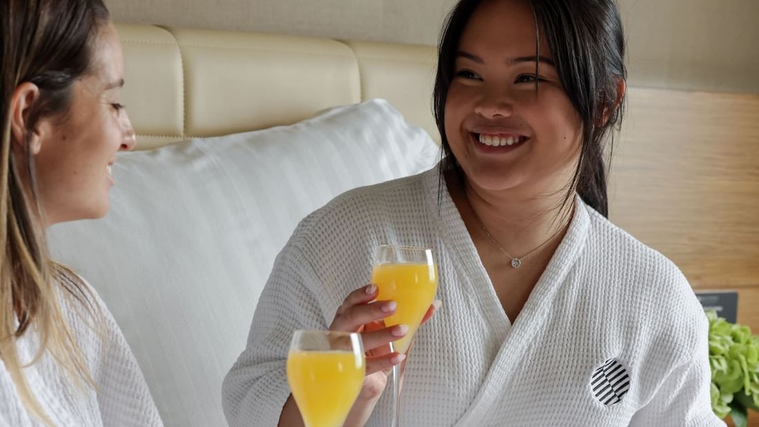 Two women in white robes toasting with glasses of orange juice in a bright hotel room with white bedding