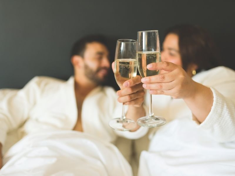 Pareja romántica en la cama de una suite brindando con champaña en Fiesta Americana Funeeq Punta Cana Hotel