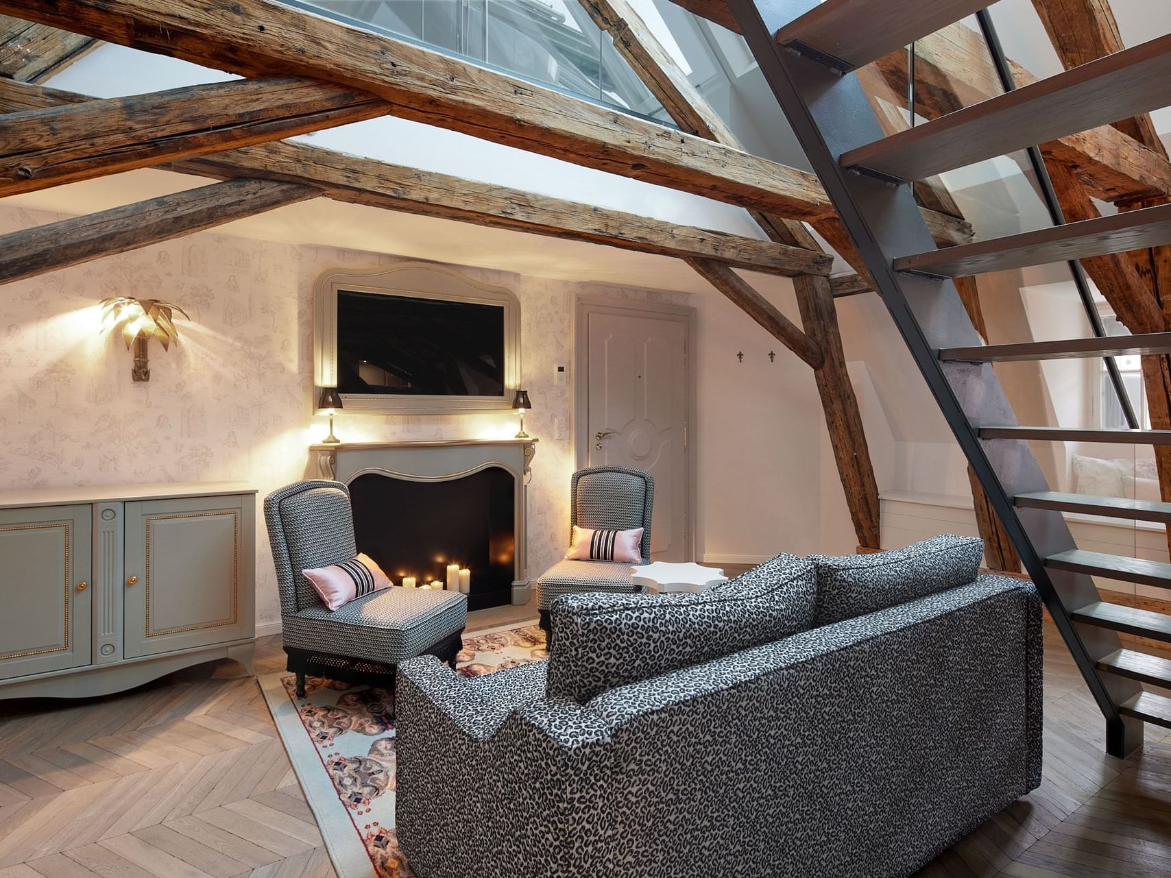 Elegant hotel room with exposed wooden beams, fireplace, couch, chairs, TV, and modern staircase.